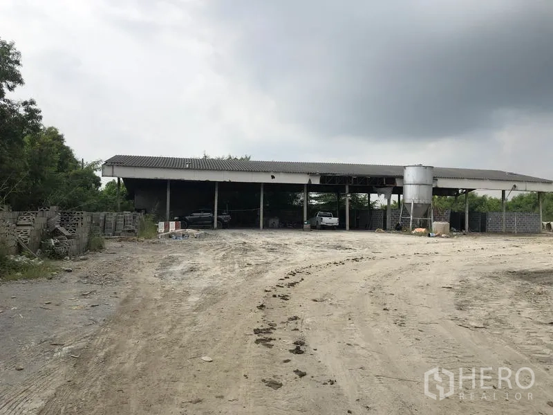 Factory for rent in Phra Nakhon Si Ayutthaya - Open shed with a vertical silo and dirt driveway, surrounded by trees and stored materials.