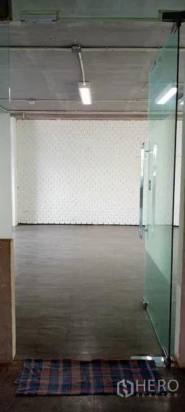 Office Space for rent in Khlong Toei, Bangkok - Glass door entry leading to an empty office featuring a white brick wall, dark flooring and linear ceiling lights.