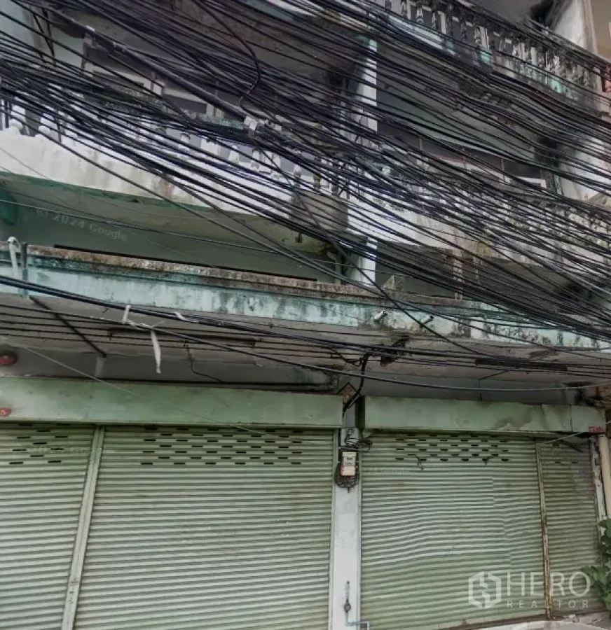 Shophouse for rent in Phra Khanong, Bangkok - Weathered multi-storey shophouse facade with closed metal roller shutters, balcony rail and dense overhead utility cables.