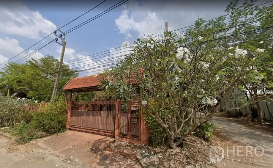 Warehouse for rent or sale in Khlong Sam Wa, Bangkok - Gated entrance with red-tiled roof and flowering trees along the boundary wall.