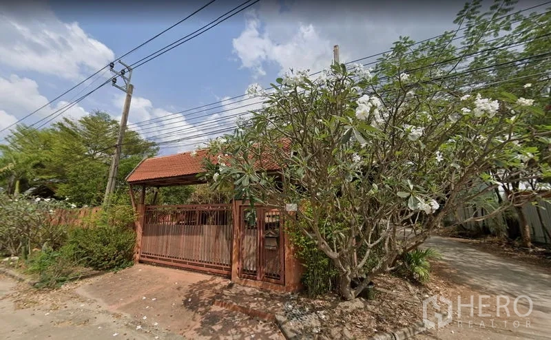 Warehouse for rent or sale in Khlong Sam Wa, Bangkok - Gated entrance with red-tiled roof and flowering trees along the boundary wall.