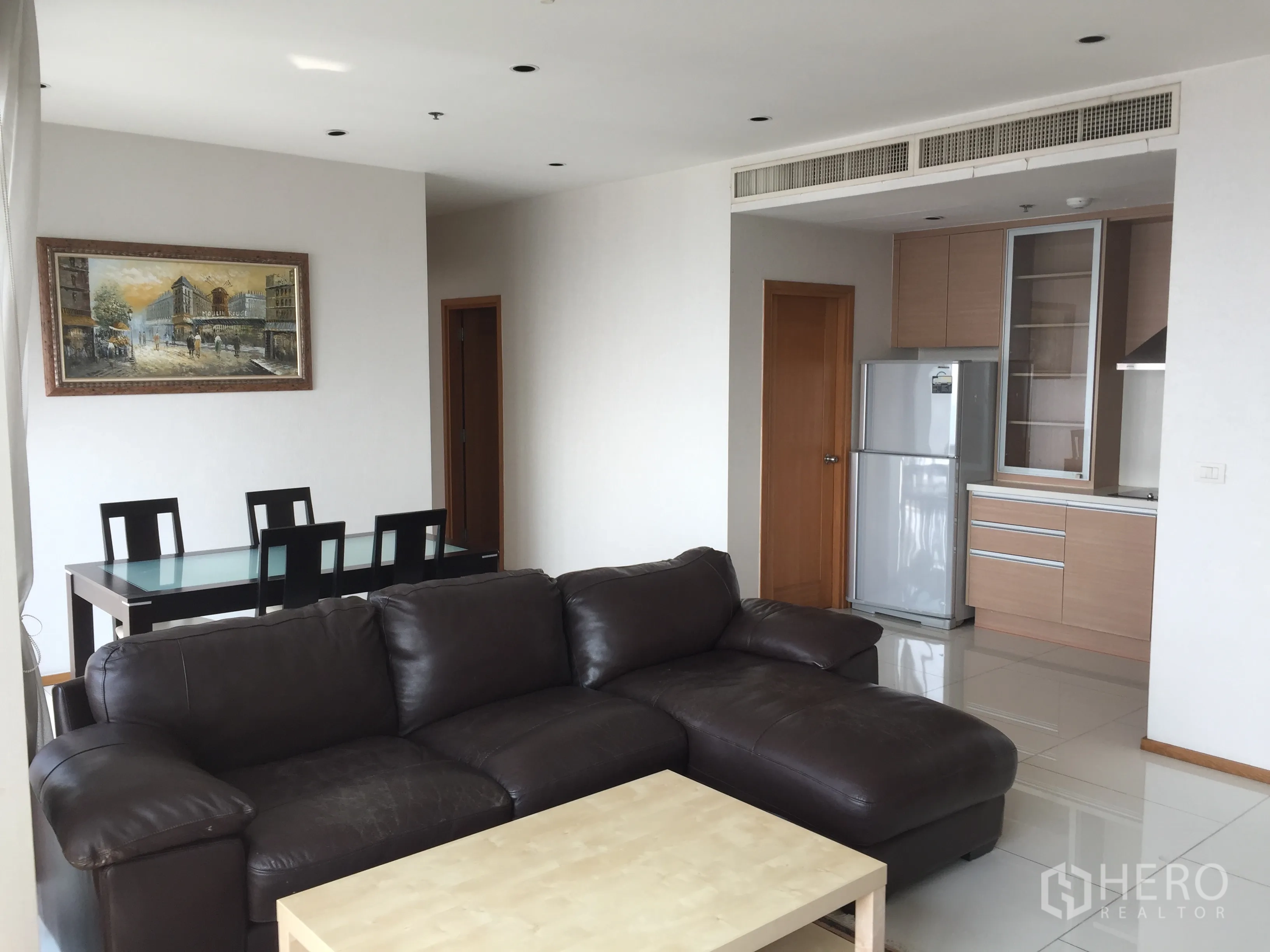 Condominium for rent or sale in Khlong Toei, Bangkok - Open-plan living room with dark leather sofa, glass dining table, and compact kitchen with stainless refrigerator.