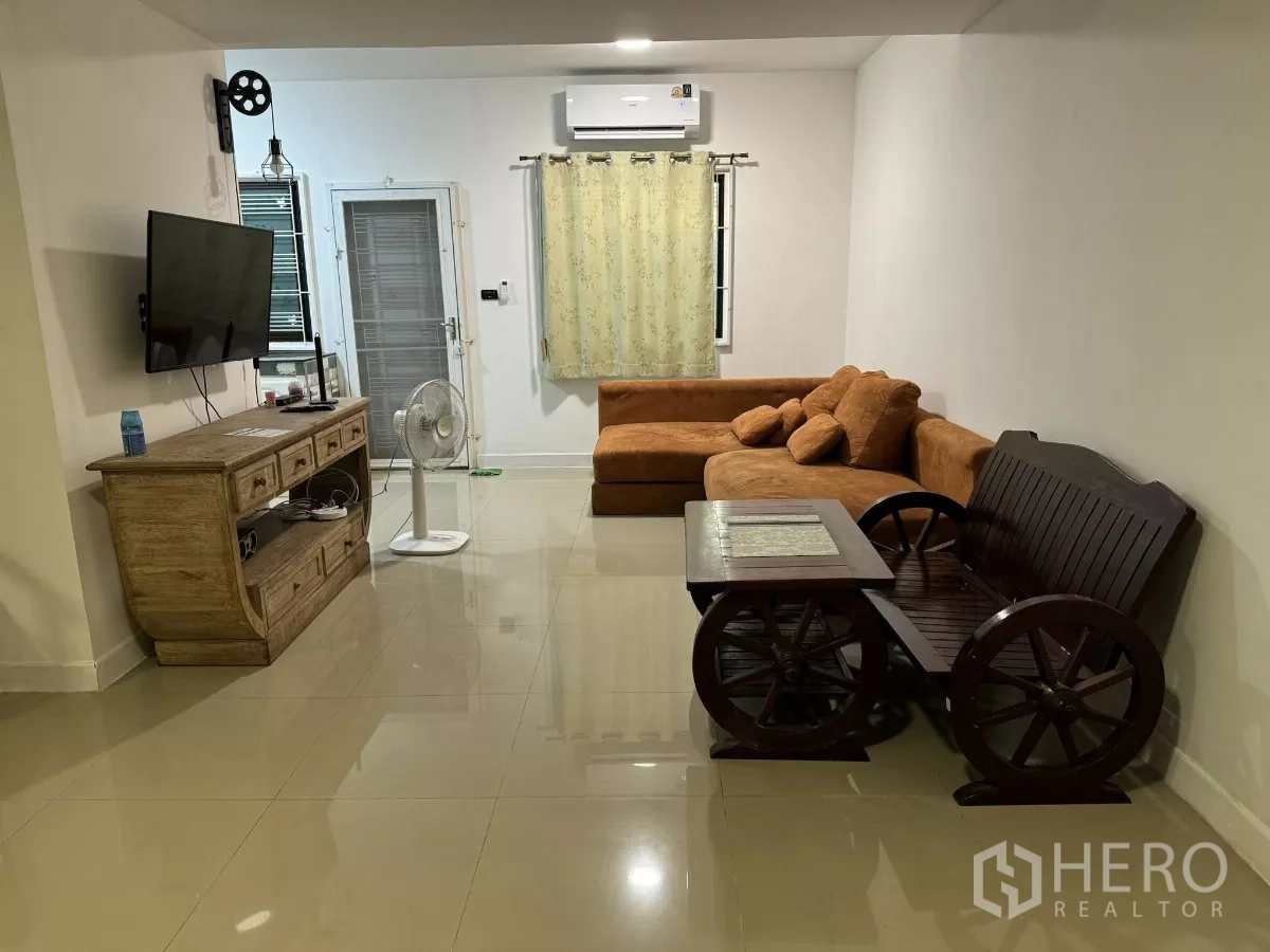 Townhouse for rent in Suan Luang, Bangkok - Air-conditioned living room with L-shaped sofa, TV console, bench, and polished tile flooring.