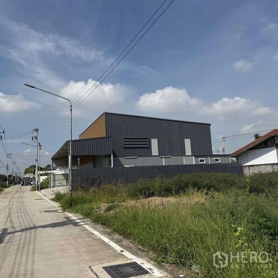 Warehouse for rent or sale in Khlong Luang, Pathum Thani - Side exterior of the warehouse along a local road with a fenced perimeter.
