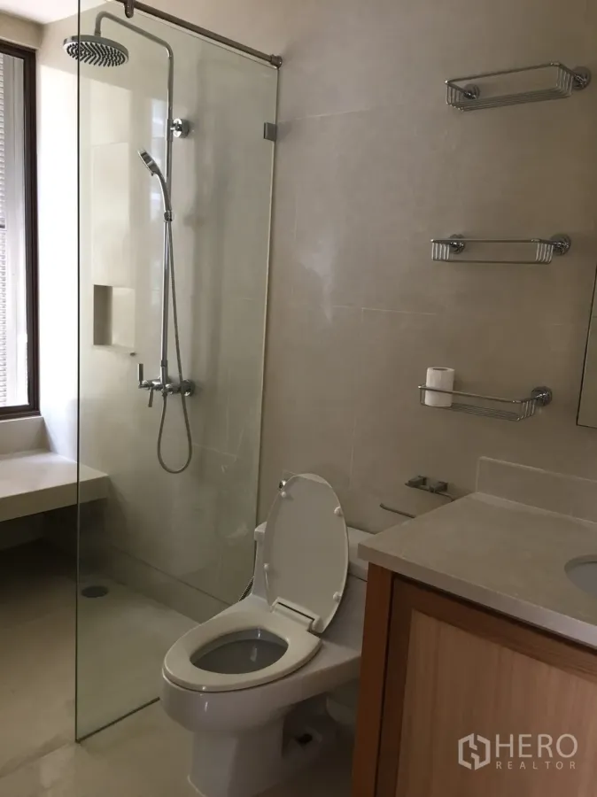 Condominium for rent in Khlong Toei, Bangkok - Modern bathroom with a glass-enclosed rain shower, wall shelves, and a light-wood vanity in a Bangkok condo.