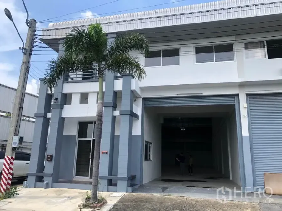 Factory for rent in Pak Kret, Nonthaburi - Modern two-storey factory facade in Pak Kret, Nonthaburi with wide roller shutter and palm tree.