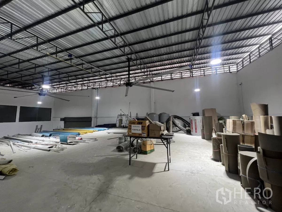 Warehouse for rent or sale in Lat Krabang, Bangkok - Spacious hall with ceiling fans, materials staged on the floor and a roller door.