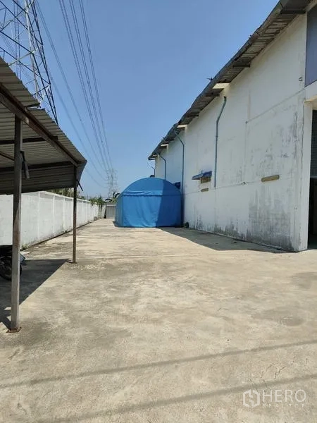 Factory for sale in Sai Noi, Nonthaburi - Large concrete yard beside the factory with a blue storage tent and overhead lines.