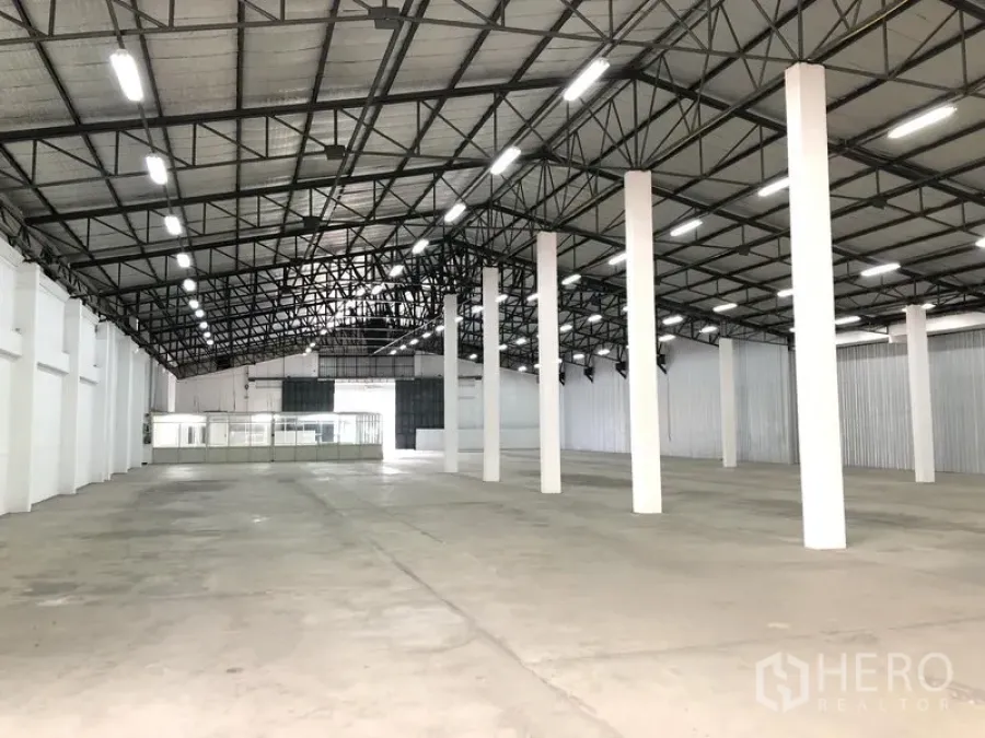 Warehouse for rent in Nonthaburi - Spacious warehouse interior with high steel-truss roof, LED lighting, white columns and concrete floor, with glass-partition office at the back.