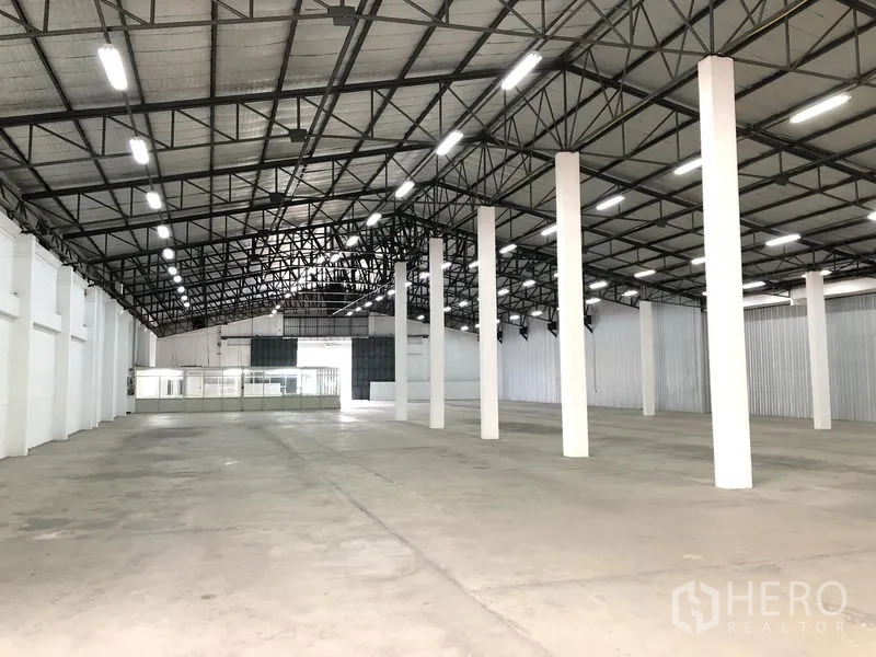 Warehouse for rent in Nonthaburi - Spacious warehouse interior with high steel-truss roof, LED lighting, white columns and concrete floor, with glass-partition office at the back.