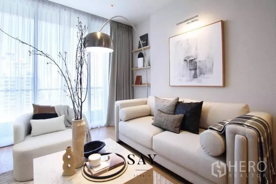 Condominium for rent or sale in Khlong Toei, Bangkok - Bright living room with cream sofa, arc floor lamp, abstract wall art and sheer floor‑to‑ceiling curtains over wood flooring.