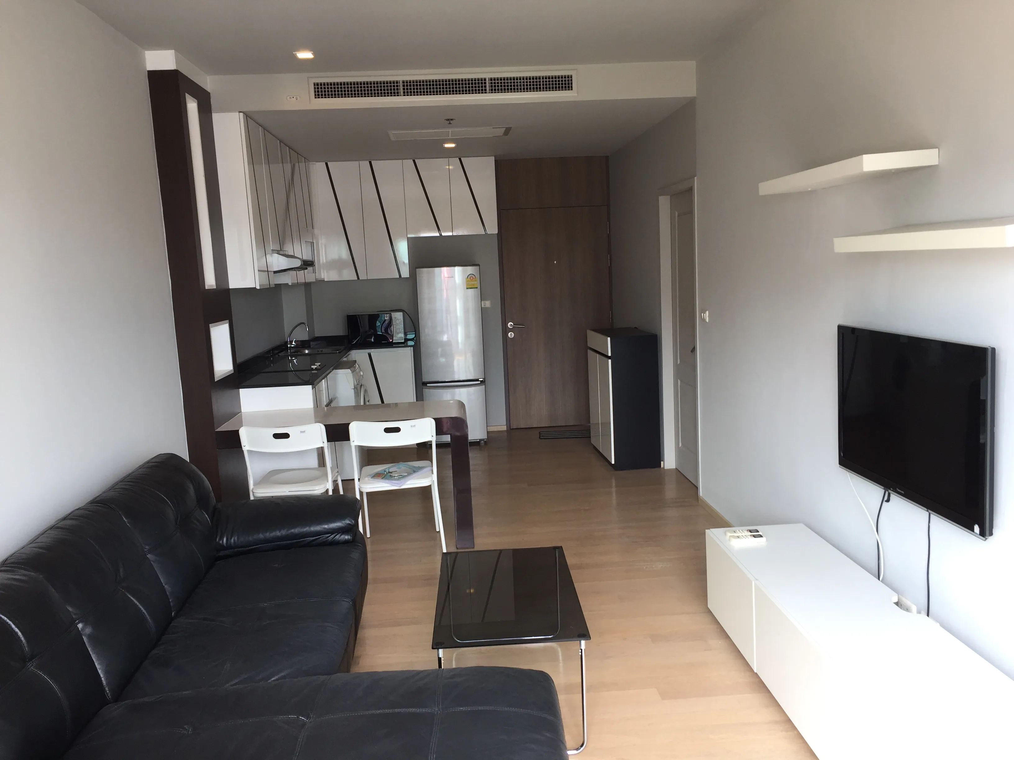 Condominium for rent in Bangkok - Modern condo interior showing living area, wall-mounted TV, dining table and sleek white kitchen cabinetry.