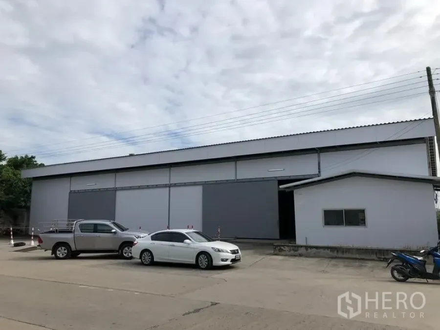 Warehouse for rent in Khlong Luang, Pathum Thani - Front exterior of a modern warehouse with large sliding doors and a wide driveway with parking space.