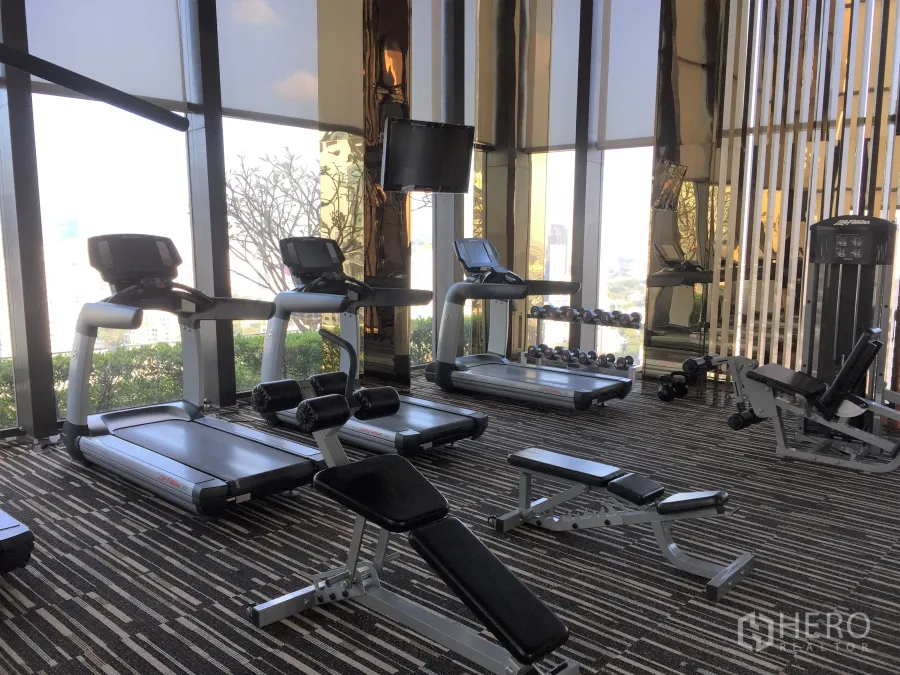 Condominium for rent or sale in Khlong Toei, Bangkok - High-rise condo fitness center with treadmills, free weights and benches facing a city view through tall windows.