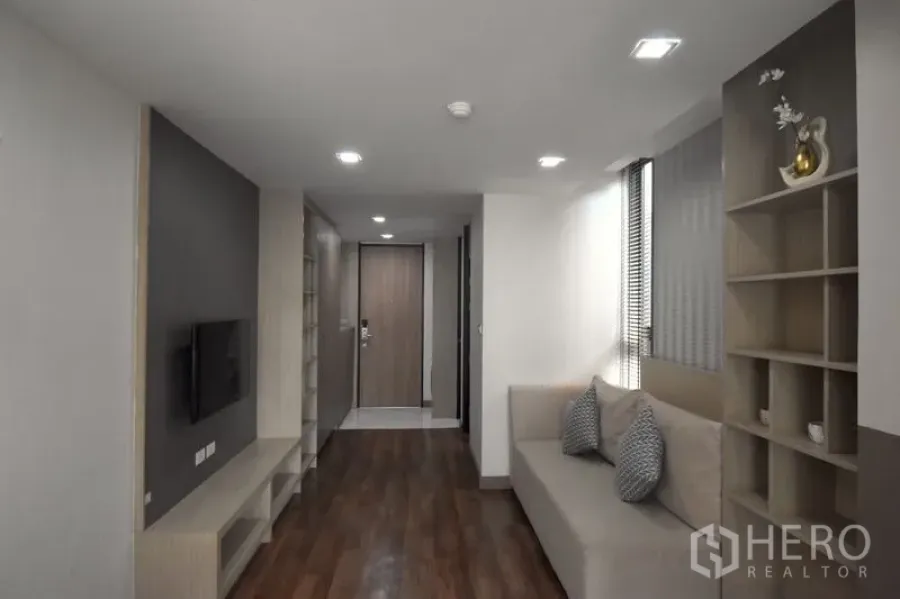 Condominium for rent in Khlong Toei, Bangkok - Living room with sofa, TV wall, wood flooring, and open display shelving.