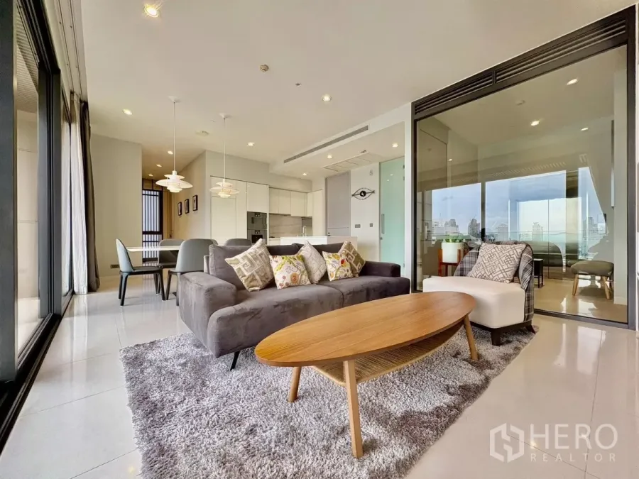 Condominium for rent in Watthana, Bangkok - Open-plan living area with gray sofa, wood coffee table, and glass-walled balcony next to modern white kitchen.