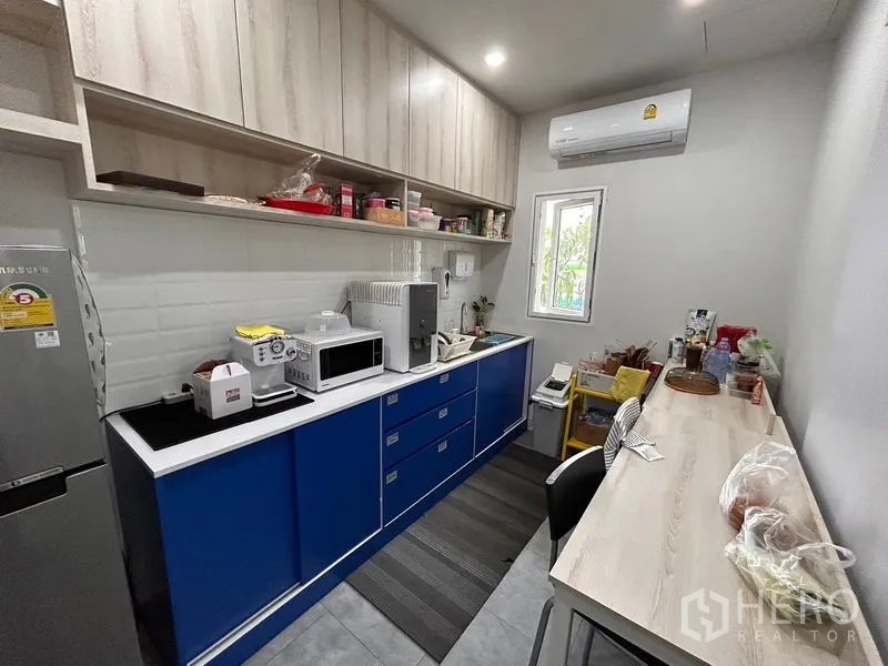 Office Space for rent in Khlong Toei, Bangkok - Pantry with blue lower cabinets, light wood overhead storage, countertop appliances and a small window with air‑conditioning.