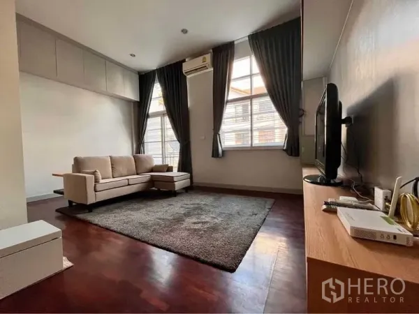 Townhouse for rent in Khlong Toei, Bangkok - Cozy living room with sofa, rug, TV console, dark curtains and wood flooring.