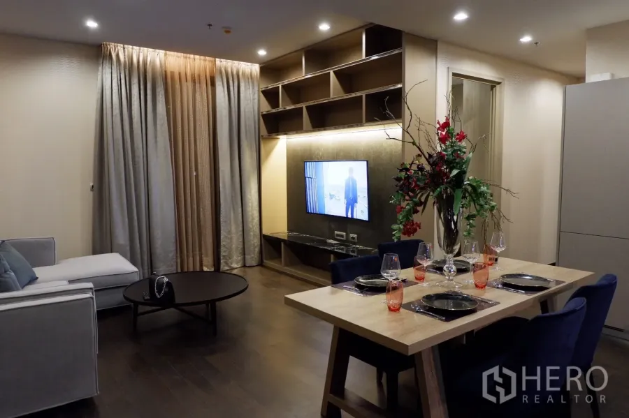 Condominium for rent in Watthana, Bangkok - Open‑plan living and dining area with built‑in TV wall, shelving, wood floors, and full‑length curtains.