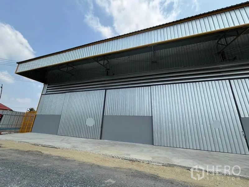 Warehouse for rent in Mueang Pathum Thani, Pathum Thani - Front exterior of a metal-clad warehouse with wide sliding doors, deep canopy and loading apron under a blue sky.