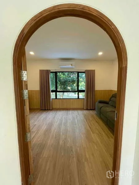 Townhouse for rent or sale in Bang Kapi, Bangkok - View through an arched door into a spacious living room with large window and curtains.