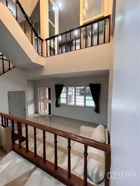 Townhouse for rent in Khlong Toei, Bangkok - Double-height living area with staircase, wooden balustrade, sofa and wide windows.
