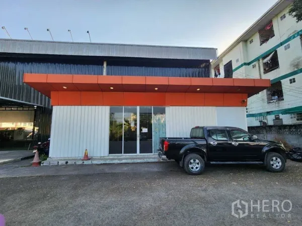Warehouse for rent in Lat Phrao, Bangkok - Front view of the glass-front office building with orange canopy and a parked pickup truck.