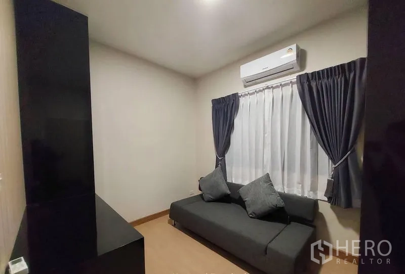 Townhouse for rent in Thanyaburi, Pathum Thani - Compact guest room with gray sofa bed, curtained window, and wall-mounted air conditioner.