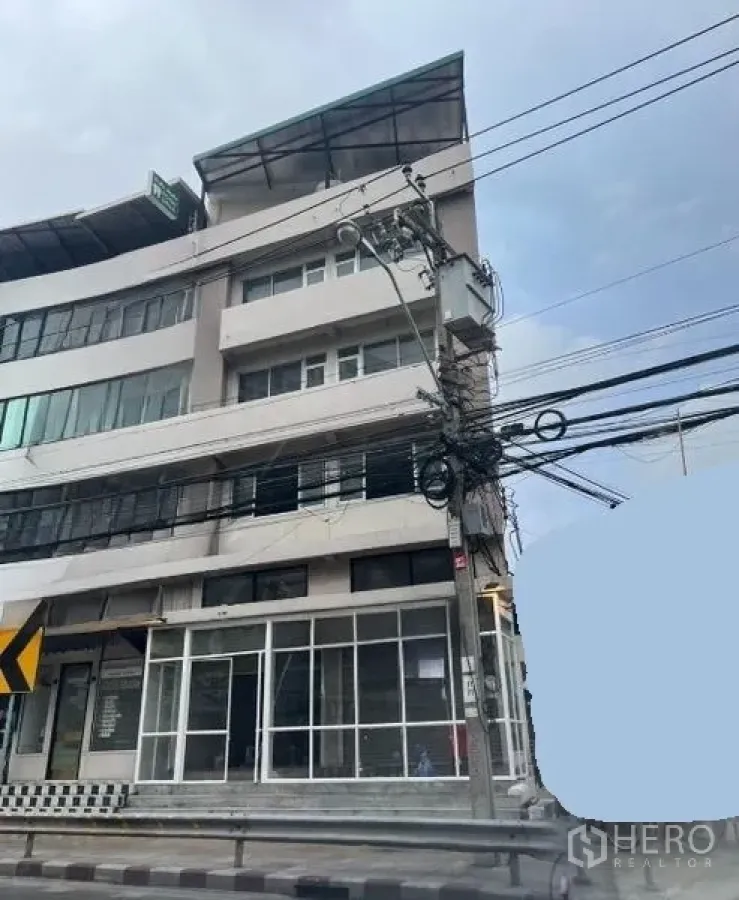Shophouse for rent in Suan Luang, Bangkok - Corner shophouse exterior showing wide windows, multiple floors, and overhead utility lines along the sidewalk.