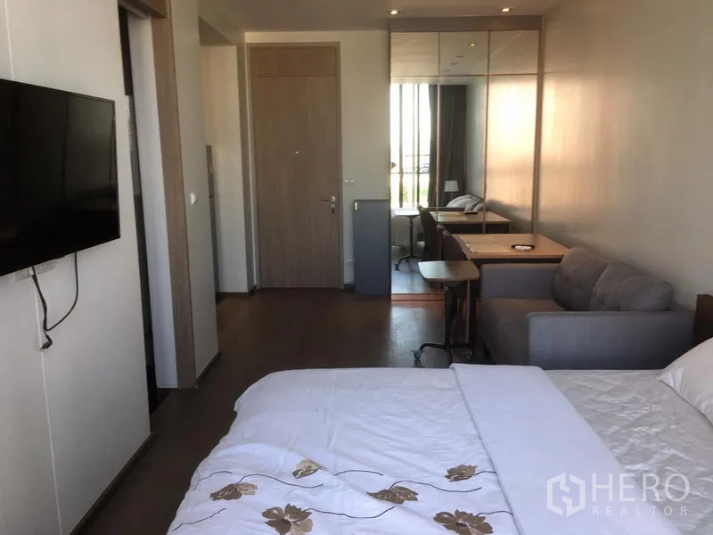 Condominium for rent in Khlong Toei, Bangkok - Bedroom view toward entrance with wall-mounted TV, work tables, sofa and mirrored wardrobe.