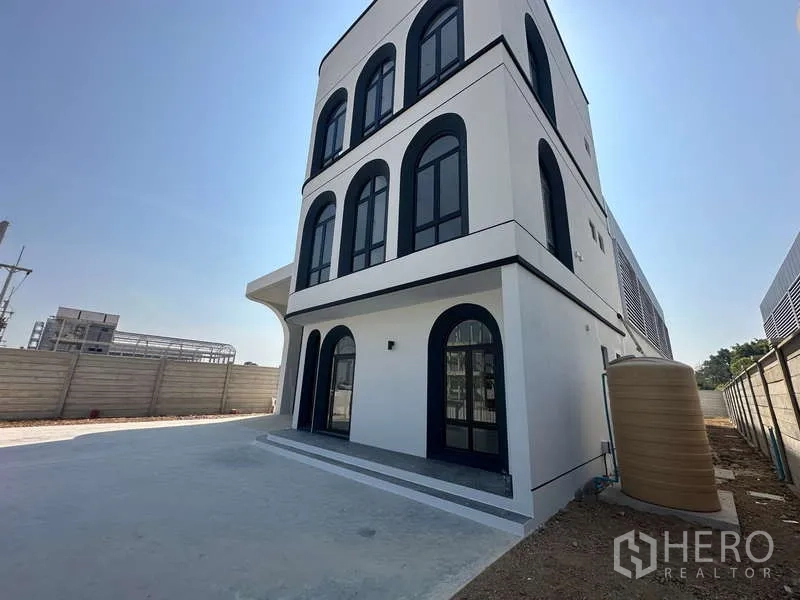 Factory for sale in Lam Luk Ka, Pathum Thani - Side view of the office building with arched windows, paved yard and a water tank by the boundary wall.