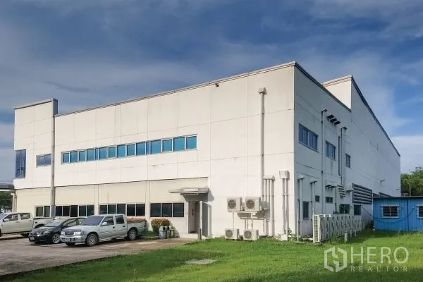 Factory for sale in Rayong - Attached office block with blue windows, air-conditioning units and parking on grass verge.