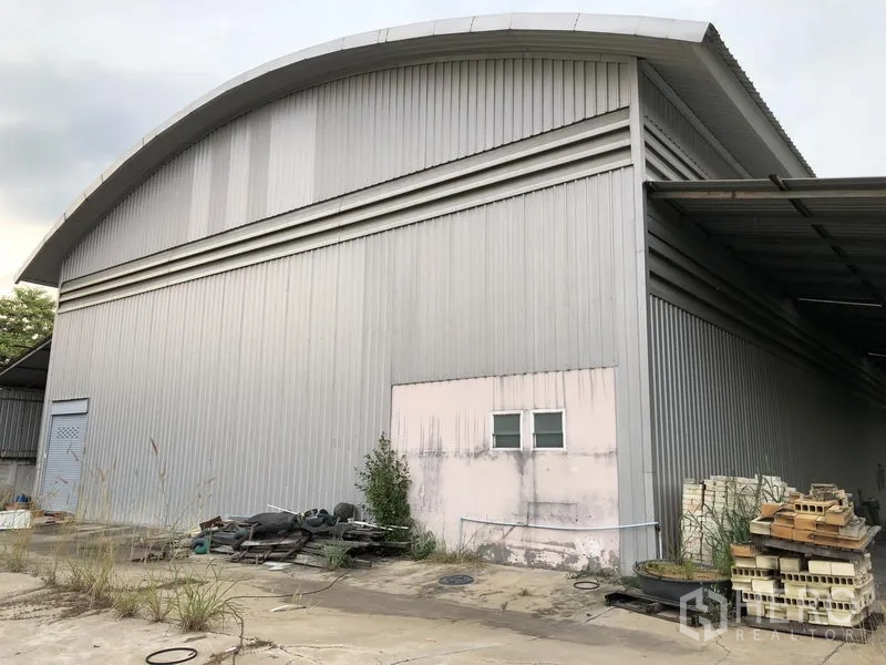 Warehouse for rent or sale in Lam Luk Ka, Pathum Thani - Side view of corrugated metal warehouse with curved roof, side canopy, service door and outdoor storage area on a concrete apron.