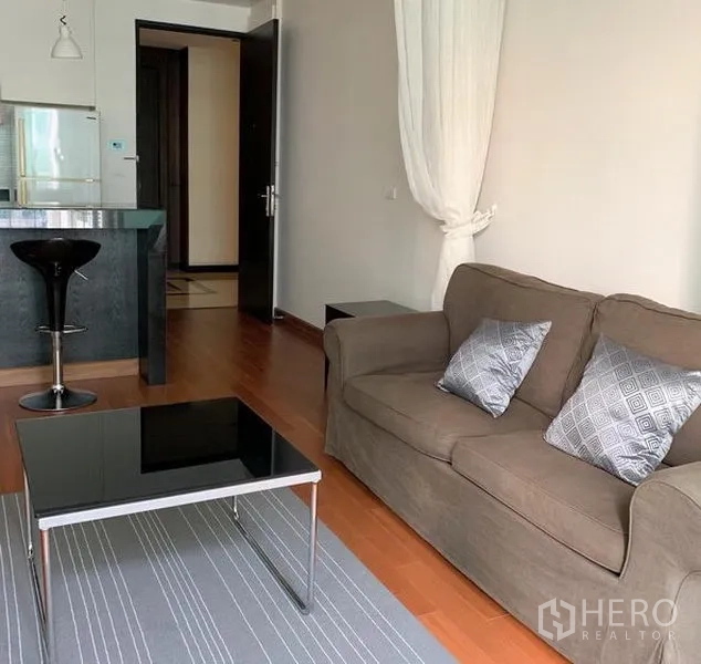 Condominium for rent in Pathum Wan, Bangkok - Bright living room with brown sofa, glass-top coffee table, and wood floors adjoining the kitchen in a Bangkok condominium.
