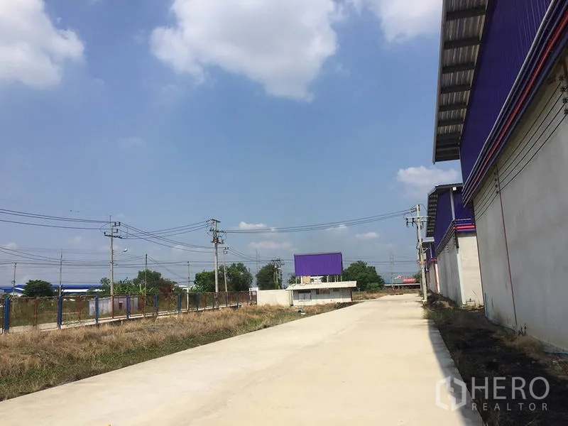 Warehouse for rent in Sai Noi, Nonthaburi - Wide concrete access road beside warehouse buildings with perimeter fencing and power lines.