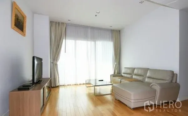 Condominium for rent in Khlong Toei, Bangkok - Bright living room with an L-shaped leather sofa, glass coffee table, TV, and floor-to-ceiling curtains.