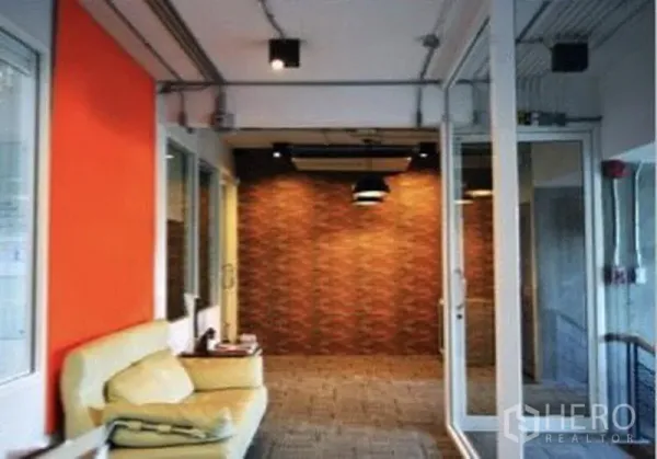 Shophouse for rent in Suan Luang, Bangkok - Upper-floor corridor with glass partitions, orange accent wall, sofa seating, exposed ceiling services, and brick feature wall.