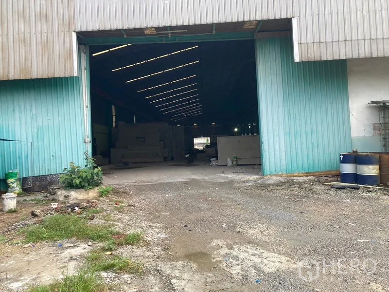 Warehouse for rent in Mueang Samut Sakhon, Samut Sakhon - Open warehouse entrance with teal doors, barrels, and a gravel loading area.