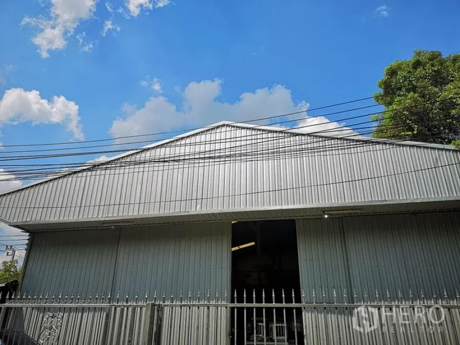 Warehouse for rent in Bang Khen, Bangkok - Front gable of metal‑clad warehouse with high roof and a partially open entrance gate.