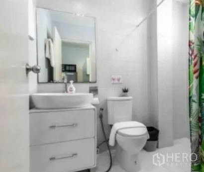 Townhouse for rent in Khlong Toei, Bangkok - Clean white bathroom with vanity, mirror, toilet, and a shower with patterned curtain.