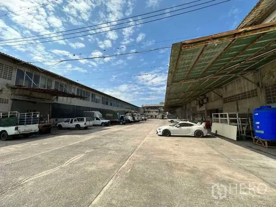 工厂 出租在Sai Noi, Nonthaburi - 工厂前方宽阔混凝土场地，设有遮雨装卸棚与卡车停车位，蓝天下景观。