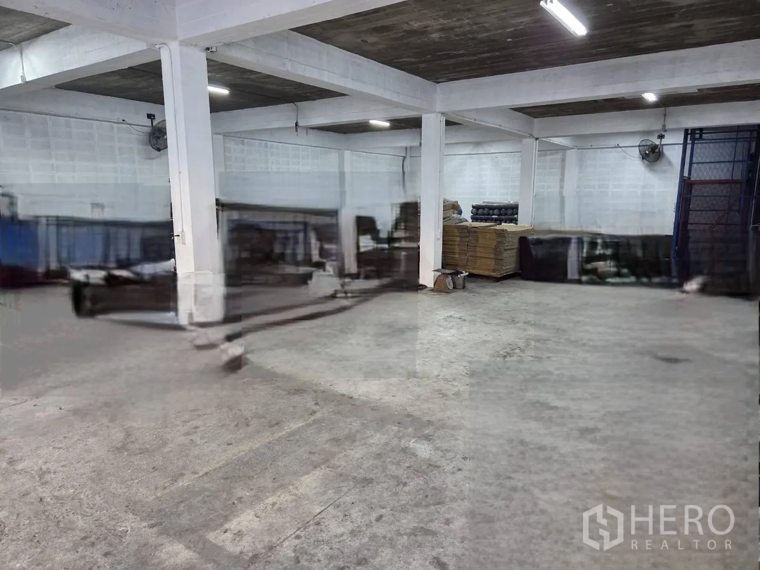 Warehouse for rent in Ratchathewi, Bangkok - Ground-floor storage area with concrete floor, structural columns, wall fans, and fluorescent lighting.