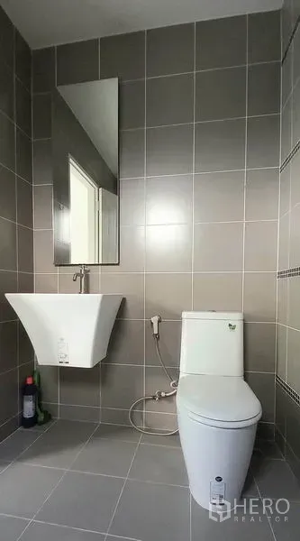 Townhouse for sale in Mueang Pathum Thani, Pathum Thani - Modern bathroom with gray tiles, wall-mounted sink, mirror, and toilet with bidet spray.