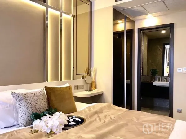 Condominium for rent in Watthana, Bangkok - Warm-lit bedroom with upholstered headboard, mirrored panels, and doorway to a marble-finished bathroom.
