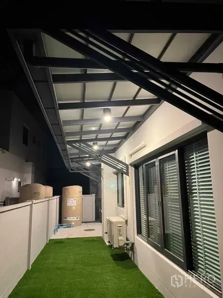 Detached House for rent or sale in Samut Prakan - Covered side patio with steel awning, artificial grass, air-con units and water tanks at night.