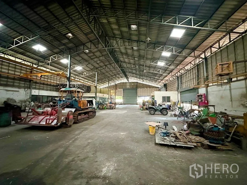 Warehouse for rent in Sai Noi, Nonthaburi - Open factory-style hall with machinery and vehicles beneath a high roof with ventilation panels.