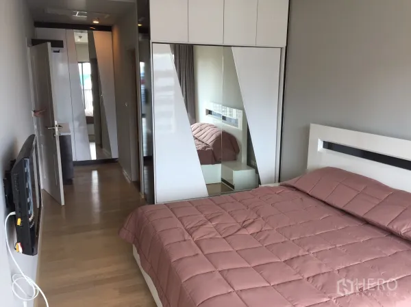 Condominium for rent in Bangkok - Bedroom featuring built-in mirrored wardrobe, wall-mounted TV and warm wood flooring.