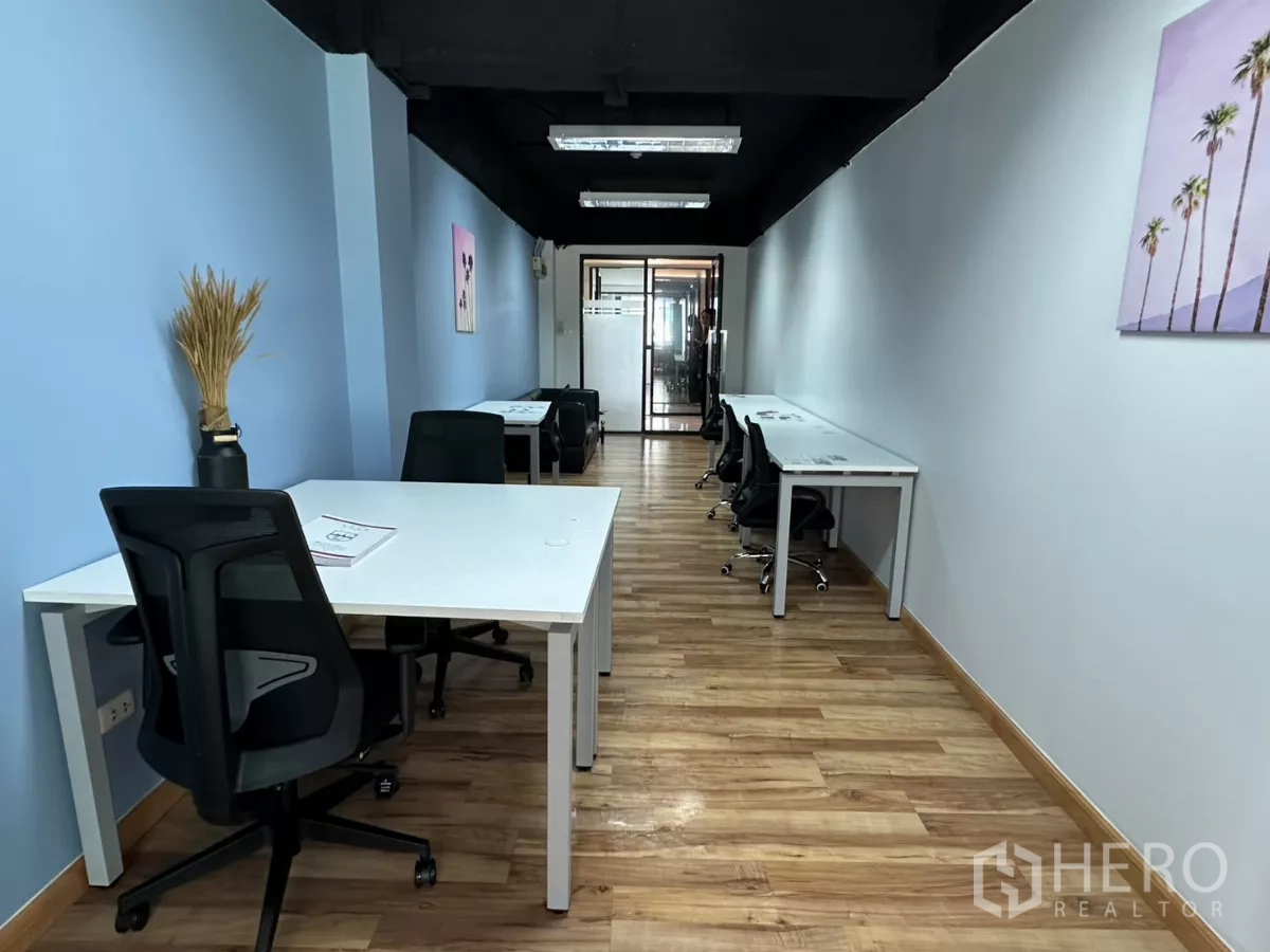 Office Space for rent in Watthana, Bangkok - Long 37 sqm furnished office with white desks, black chairs, blue walls, wood floors and fluorescent lights leading to a glass meeting room.