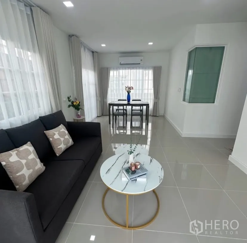 Detached House for rent in Mueang Samut Prakan, Samut Prakan - Living room with dark sofa, round coffee table and dining set by large windows.