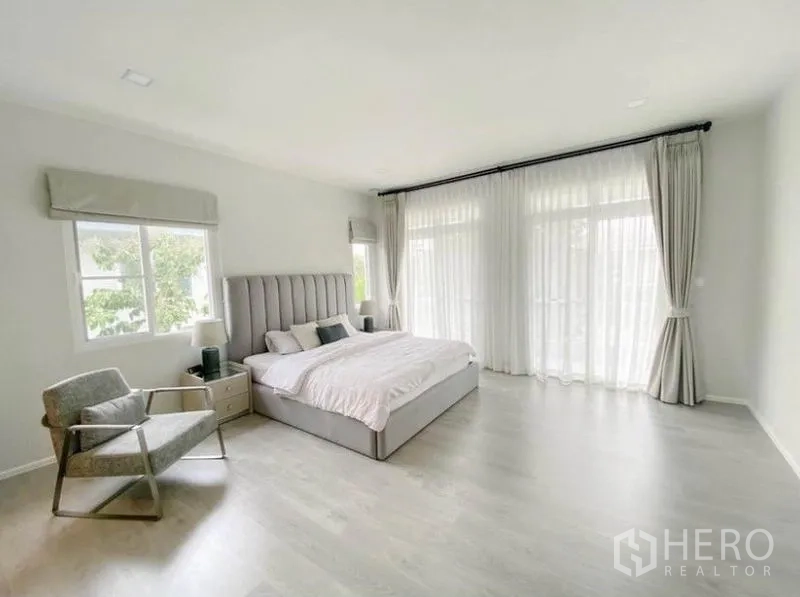 Detached House for rent in Prawet, Bangkok - Spacious primary bedroom with king bed, lounge chair and wall-to-wall curtains to the balcony.